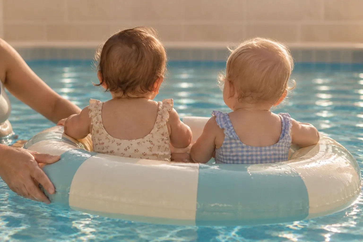 Zwei Babys sitzen von hinten gesehen in einem hellblau-weißen Schwimmring im Becken, eine Hand hält den Ring